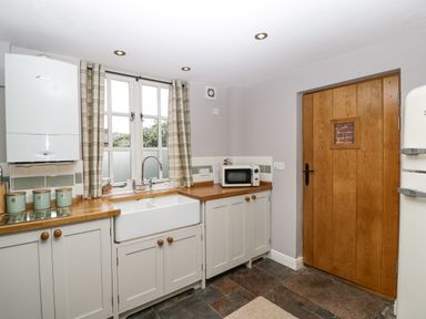 A kitchen with white cabinets wooden countertops a white sink a window with curtains a microwave and a wooden door at The Old Bakehouse in Pembridge