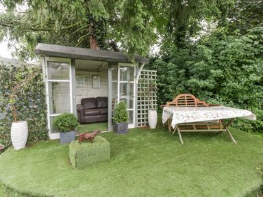 A garden with a small wooden shed containing a sofa next to a table with a floral tablecloth and a bench at The Old Bakehouse in Pembridge