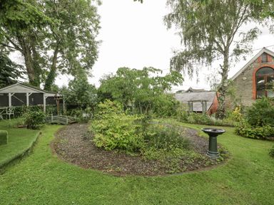 A garden with a circular flower bed a bird bath a small wooden bridge and a stone building at The Old Bakehouse in Pembridge