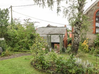 A garden with shrubs flowers a tree and a small stone and brick building at The Old Bakehouse in Pembridge