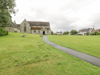 A stone church with a paved path leading to its entrance and a grassy field with benches and houses in the background at The Old Bakehouse in Pembridge