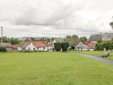 A green open field with a paved path leading to a cluster of houses under a cloudy sky at The Old Bakehouse in Pembridge