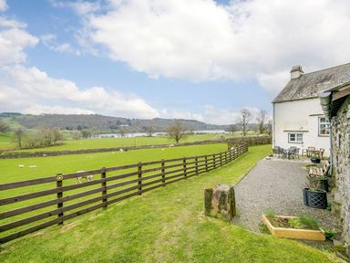 A grassy yard with wooden fence and outdoor seating next to a stone and white house overlooking fields and a lake at Shepherds Cottage in Hawkshead