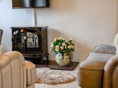 A black stove heater with a vase of white flowers next to it and beige armchairs in a living room at Shepherds Cottage in Hawkshead
