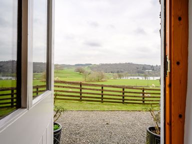 A view through an open door showing a gravel path a wooden fence green fields trees and a lake at Shepherds Cottage in Hawkshead
