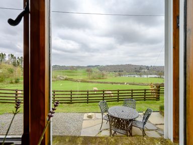 A view through a window of a patio with metal table and chairs and a green field with sheep at Shepherds Cottage in Hawkshead