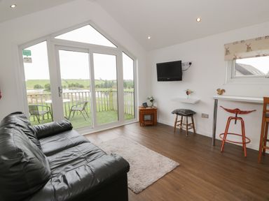 A living room with a black leather sofa a wall mounted TV a small wooden nesting table and stools near large windows overlooking a balcony with chairs at Mallard Lodge in Moscow near Galston