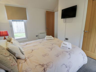 A bedroom with a bed covered in patterned bedding two stacks of white towels a window with a closed blind a wall-mounted tv and two wooden doors at Mallard Lodge in Moscow near Galston