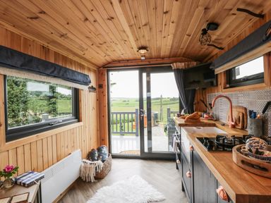 A kitchen with wooden walls and ceiling a countertop with a sink stove and cutting board and sliding glass doors showing a green landscape at Greenacres Carriage in Maybole