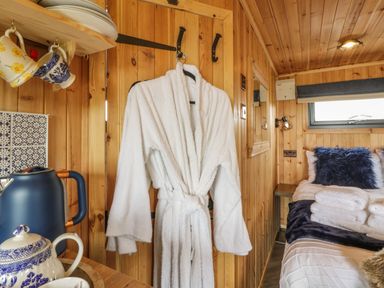 A wooden room with a white bathrobe hanging on hooks a bed with pillows and folded towels and kitchenware on shelves at Greenacres Carriage in Maybole