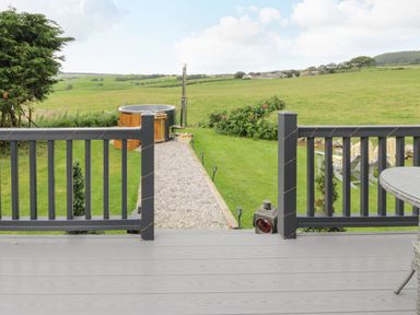 An outdoor deck with a railing overlooking a gravel path leading to a wooden hot tub and grassy field at Greenacres Carriage in Maybole
