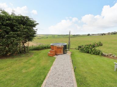 A wooden hot tub on a gravel path in a grassy backyard with bushes and fields at Greenacres Carriage in Maybole