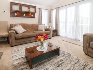 A living room with a brown sofa and pillows a wooden coffee table with flowers a side table with a lamp and curtains at Gwel Eryri in Penmynydd near Llanfairpwllgwyngyll