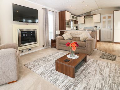 A living room with a sofa armchair coffee table with flowers wall-mounted tv and fireplace with an open kitchen in the background at Gwel Eryri in Penmynydd near Llanfairpwllgwyngyll