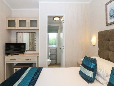 A bedroom with a bed, two blue pillows, a desk with a television and a mirror, and a doorway leading to a bathroom at Gwel Eryri in Penmynydd near Llanfairpwllgwyngyll