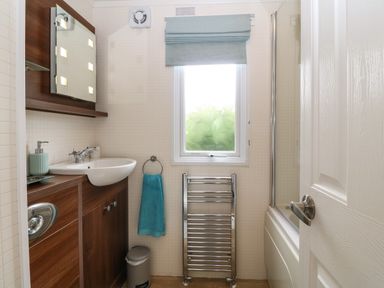 A small bathroom with a sink and wooden cabinet a window with a blue blind a chrome heated towel rail a blue hand towel and a shower behind a white door at Gwel Eryri in Penmynydd near Llanfairpwllgwyngyll