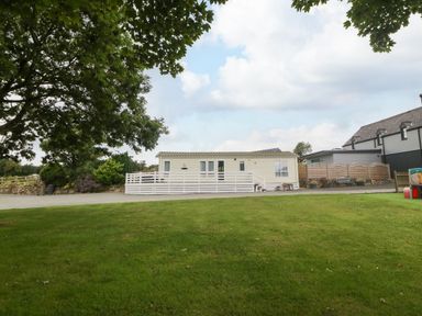 A single story mobile home with a white fenced porch on a grassy plot with a large tree nearby at Gwel Eryri in Penmynydd near Llanfairpwllgwyngyll