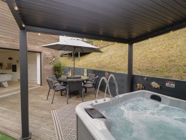 A covered outdoor patio with a hot tub and dining table under an umbrella at 12 Beachdown in Challaborough