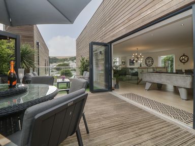 An outdoor patio with a glass table and chairs opening into a room with a pool table and modern lighting at 12 Beachdown in Challaborough