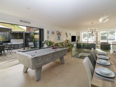 A game room with a pool table dining table set and sofas with large windows and an outdoor seating area at 12 Beachdown in Challaborough