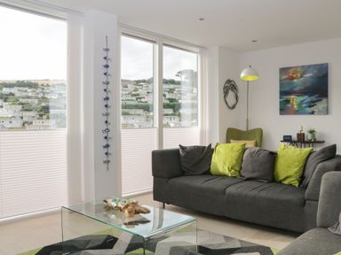 A living room with a gray sofa and green pillows a glass coffee table and large windows at 12 Beachdown in Challaborough