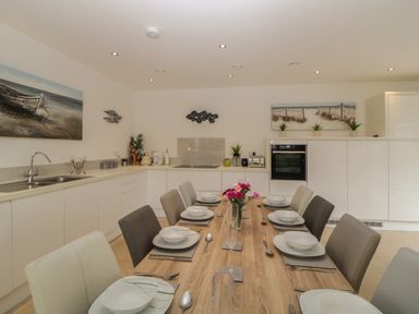 A kitchen and dining area with a wooden dining table set with white dishes and cutlery and white and gray chairs at 12 Beachdown in Challaborough