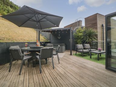 An outdoor patio with a round table and six chairs under a large umbrella and additional seating near plants at 12 Beachdown in Challaborough