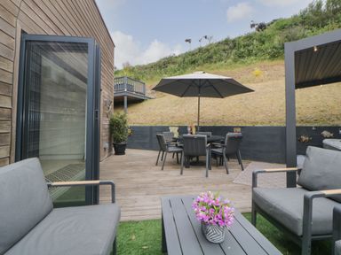 An outdoor patio area with grey cushioned seating, a table with pink flowers, a dining table with chairs and an umbrella at 12 Beachdown in Challaborough