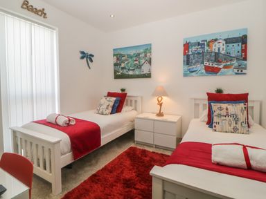 A bedroom with two single beds with red throws and cushions a white nightstand with a lamp and two paintings on the wall at 12 Beachdown in Challaborough