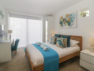 A bedroom with a bed with blue and sea-themed pillows a desk with a chair and lamp and vertical blinds at 12 Beachdown in Challaborough
