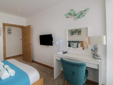 A bedroom with a wooden bed, a wall-mounted television, a white desk with a mirror, fan, lamp, and teal chair at 12 Beachdown in Challaborough