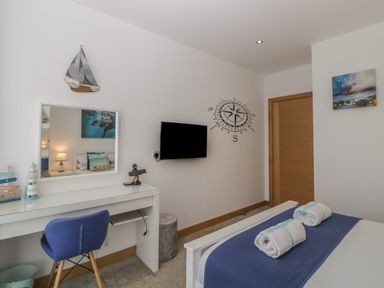 A bedroom with a blue chair a white desk a wall-mounted television and a bed with rolled towels at 12 Beachdown in Challaborough