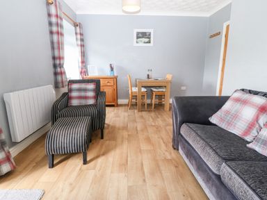A living room with a sofa and armchair with footstool a wooden dining table with four chairs and a wooden sideboard at Coed y Glyn Bach Equestrian in Penygroes