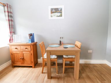 A dining area with a wooden table and two chairs beside a wooden cabinet and a window with curtains at Coed y Glyn Bach Equestrian in Penygroes