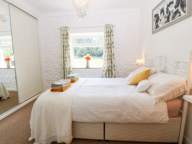 A bedroom with a bed with white bedding and yellow and white pillows next to a window with floral curtains and a mirrored wardrobe at Coed y Glyn Bach Equestrian in Penygroes