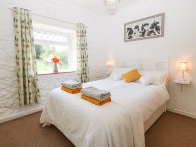 A bedroom with a double bed white bedding and cushions two lamps and a window with curtains and flowers at Coed y Glyn Bach Equestrian in Penygroes