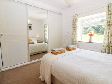A bedroom with a bed covered in white bedding folded towels on top a window with floral curtains and a vase of flowers on the windowsill a large wardrobe with mirrored sliding doors