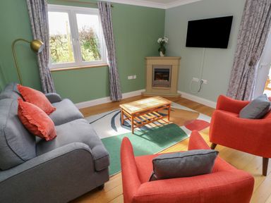 A living room with a gray sofa and orange chairs a wooden coffee table a wall-mounted TV and a corner fireplace at The Annexe Meadow Croft in Haverfordwest