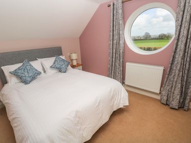 A bedroom with a double bed white bedding two blue pillows a bedside table lamp round window with curtains and a view of a field at The Annexe Meadow Croft in Haverfordwest