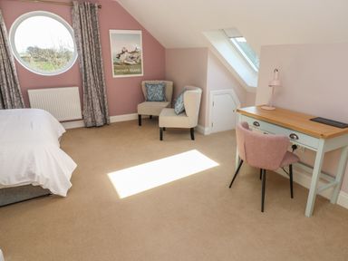 A bedroom with a bed, round window with curtains, two chairs with cushions, a desk with a lamp and chair at The Annexe Meadow Croft in Haverfordwest