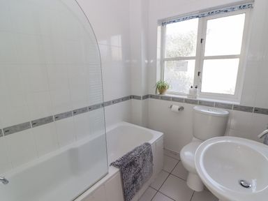 A bathroom with a bathtub with a glass shower screen and towel a toilet and a sink under a window with a plant at 3 Strawberry Close in Little Haven near Broad Haven