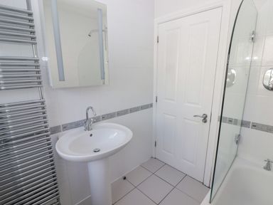 A bathroom with a white sink and pedestal a mirror a heated towel rail a bathtub with a glass shower screen and a white door at 3 Strawberry Close Little Haven near Broad Haven