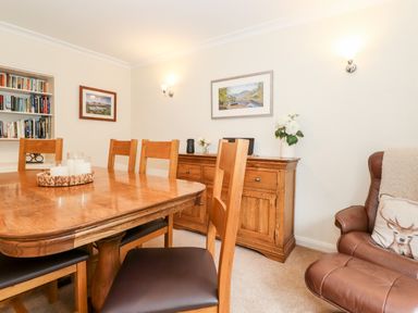 A dining room with wooden table and chairs a wooden sideboard with decorations built-in bookshelf and a leather armchair with a cushion at Holly Cottage in Grasmere