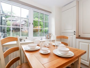 A dining area with a wooden table set with white dishes and cups near a window with white frames at Holly Cottage in Grasmere