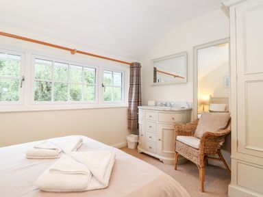 A bedroom with a bed, white towels, a wicker chair with cushions, a white cabinet with a mirror, and windows with curtains at Holly Cottage in Grasmere