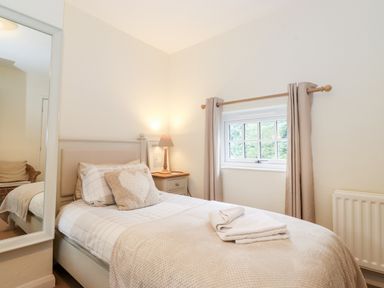 A bedroom with a single bed a bedside table with a lamp a window with beige curtains and a large mirror on the wall at Holly Cottage in Grasmere