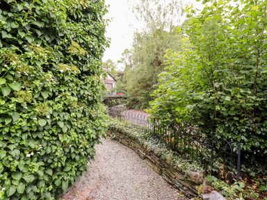 A gravel path bordered by a stone wall and metal fence with dense green foliage and trees near a river at Holly Cottage in Grasmere