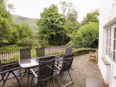 An outdoor patio area with a glass table and six black chairs beside a white building at Holly Cottage in Grasmere