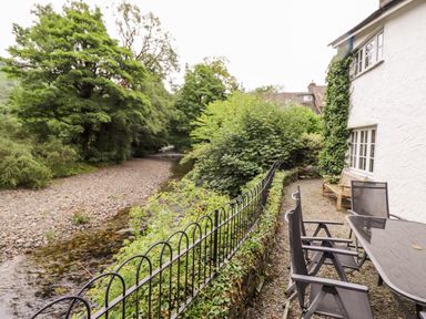 An outdoor seating area with chairs and a table next to a white building by a river with trees and bushes at Holly Cottage in Grasmere