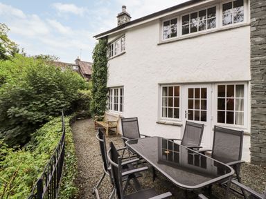 An outdoor patio area with a glass table, six chairs, a wooden bench, and white building walls at Holly Cottage in Grasmere